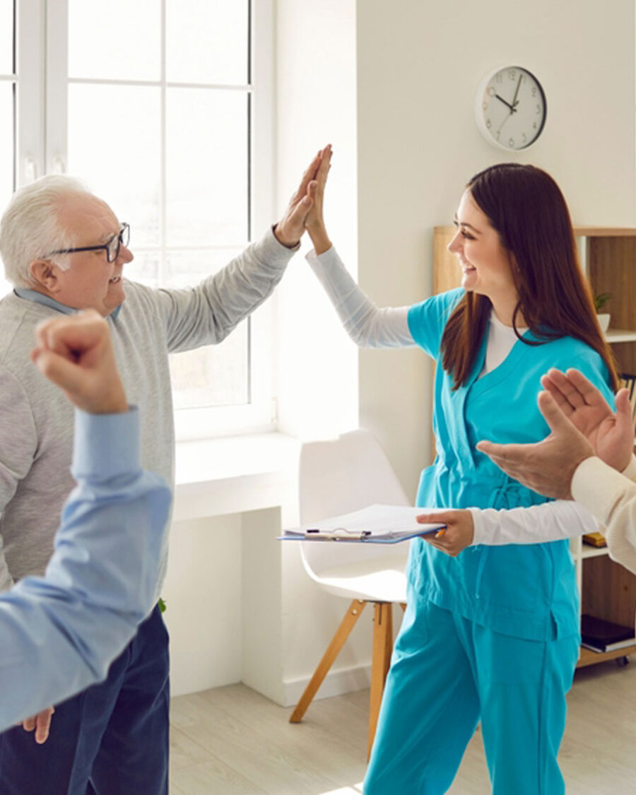 Pflegerin gymnastiziert mit Senior:innen in einem Pflegeheim. Mit einem der Senioren macht sie einen "High Five"-Abschlag, Brain City Berlin