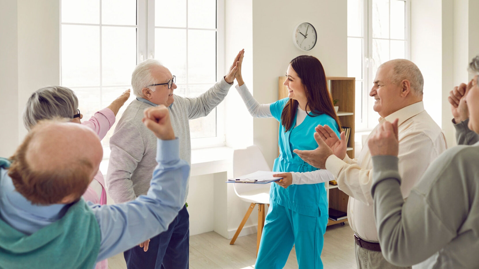 Pflegerin gymnastiziert mit Senior:innen in einem Pflegeheim. Mit einem der Senioren macht sie einen "High Five"-Abschlag, Brain City Berlin