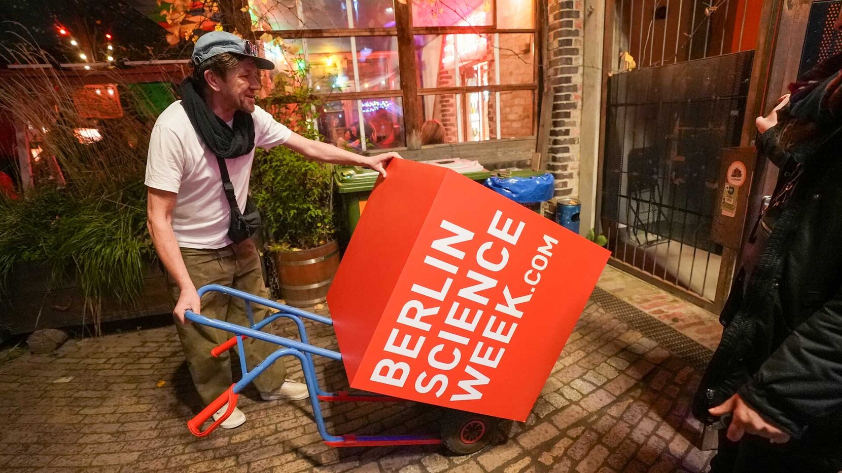 Mann mit Schubkarre schiebt roten Logo-Würfel der Berlin Science Week, Brain City Berlin