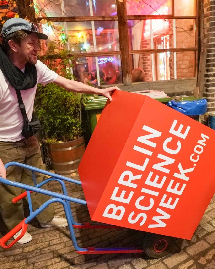 Mann mit Schubkarre schiebt roten Logo-Würfel der Berlin Science Week, Brain City Berlin